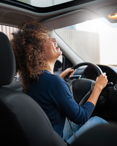 Eine Frau sitzt hinter dem Steuer eines Autos, nimmt den Kopf in den Nacken und lacht herzlich.