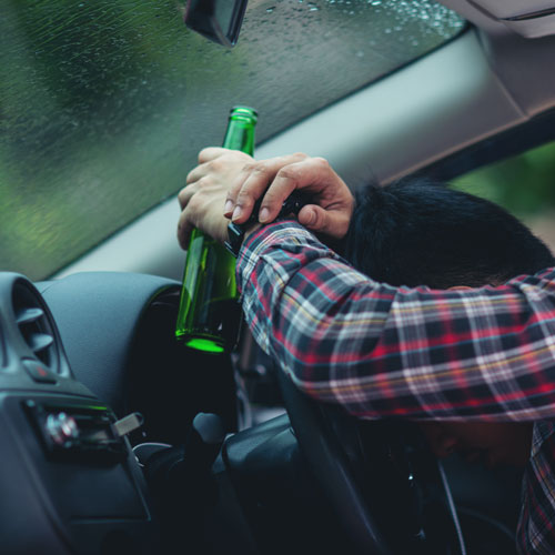 Ein Mann sitzt am Steuer seines Autos mit dem Kopf auf dem Lenkrad und einer Bierflasche in der Hand.